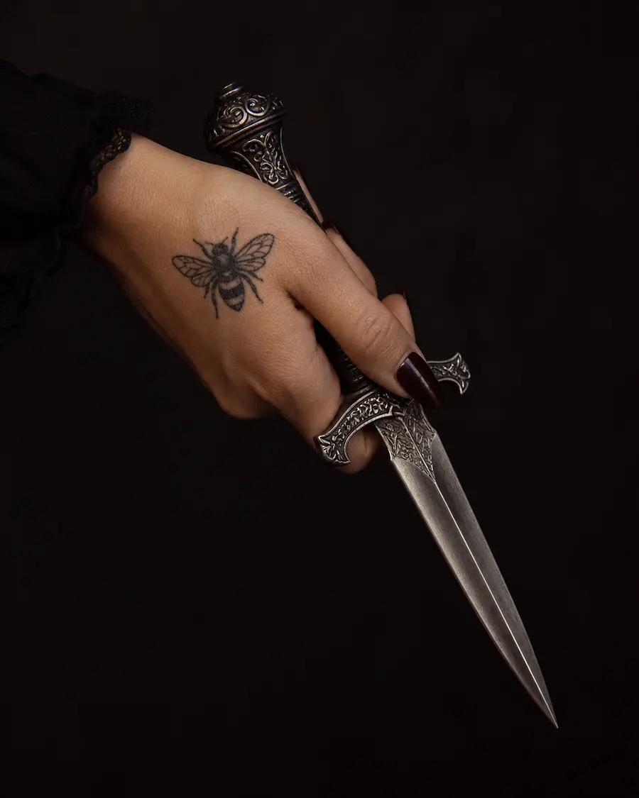 Woman’s hand, holding a dagger, with a bee tattoo on the hand