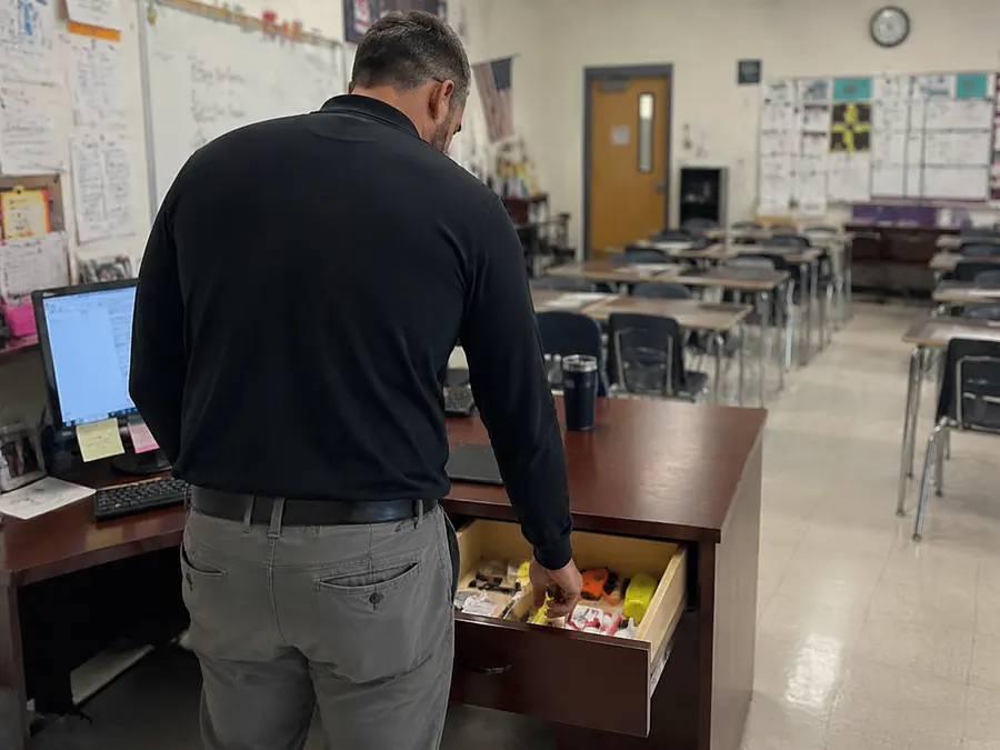 Male teacher standing at his desk putting his finger into an open desk drawer...