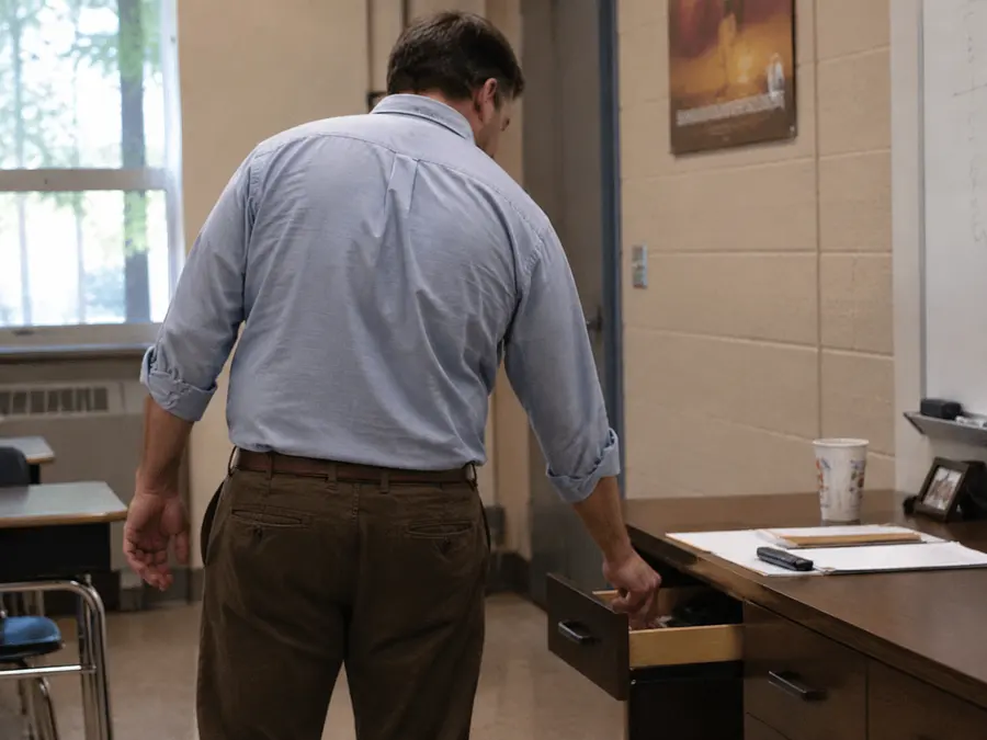 Male teacher standing at his desk putting his finger into an open desk drawer...